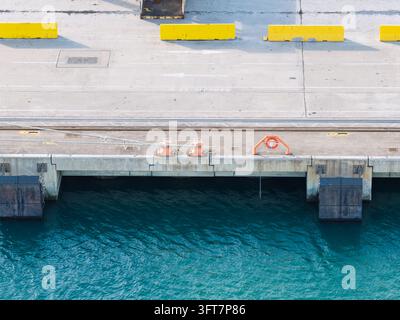 Luftaufnahme eines Betonpiers mit gelben Sicherheitsbarrieren, Festmacherpollern und einer Rettungsboje entlang des Wasserrandes im Vereinigten Königreich. Stockfoto