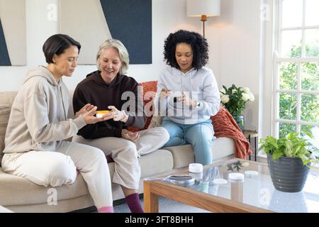 Verschiedene Freundinnen in Kapuzenpullovern untersuchen kleine Gläser auf einem Glas-Couchtisch im Wohnzimmer Stockfoto
