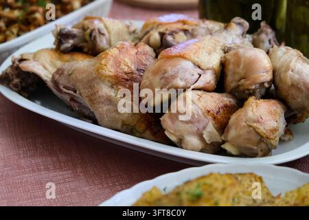 Gebratene Hühnerstücke und gewürzte Kartoffelkeile, serviert auf weißen Keramiktellern, mit Gläsern mit eingelegten Gurken im Hintergrund. Stockfoto