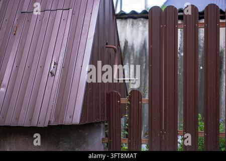 Detailaufnahme einer lila-lilawelligen Metalldachkante mit Messingbeschlägen, angrenzender dunkelbrauner Holzlattenzaun mit Metallverschluss. Stockfoto