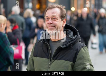 Ein Mann mittleren Alters lächelt auf einer belebten Straße, während Passanten im Hintergrund verschwommen sind. Stockfoto