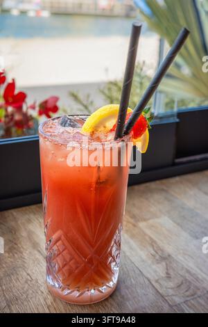 Erdbeerlimonade-Geberdrink mit zwei Papierstrohhalmen und einer Zitronenscheibe in einem hohen Glas auf einem Holztisch am Fenster. Stockfoto
