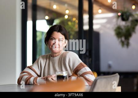 Frau sitzt an einem Tisch in einem gemütlichen Café, lächelt neben einem Glas dunklem Kaffee, trägt einen gestreiften Pullover Stockfoto