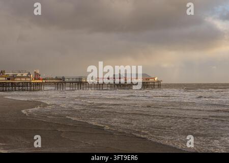 Wellen spülen über nassen Sand unter der Central Pier von Blackpool, wobei Pier-Gebäude und Metallstützen von warmem Sonnenlicht unter grauen Wolken beleuchtet werden. Stockfoto