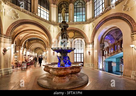 Der Donaudonauf-Brunnen leuchtet blau im Innenhof der historischen Einkaufspassage, während Fußgänger an einem Café an einem Herbstabend in Österreich vorbeigehen Stockfoto