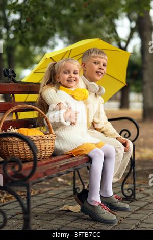 Kindheitsfreundschaft und Herbstsaison, glücklicher Bruder und Schwester sitzen auf einer Parkbank mit gelbem Regenschirm und Kürbis im Korb an einem Herbsttag Stockfoto