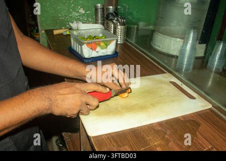 Männerhände, die mit einem Messer eine orangefarbene scharfe Paprika auf einem Schneidebrett schneiden. Kommerzielle Küchenzubereitung zeigt das Würfeln von heißen aromatischen Zutaten. Stockfoto