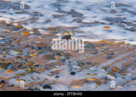 Ansicht von nassen Kieselsteinen und glatten Steinen auf dem Sandstrand mit weißem Seeschäum von anrollenden Wellen in Talacre, Flintshire, Vereinigtes Königreich. Stockfoto