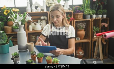Floristin in Schürze tippt auf ein Tablet an der Blumenladentheke zwischen Topfpflanzen, Vase und Geschenkpapier; ruhige Konzentration. Stockfoto