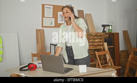 Frauenschreinerin, die ein Smartphone ans Ohr hält und sich auf einen Tisch mit Laptop und roten Ohrschützern lehnt, in einem Studioarbeitsplatz; praktische Sorge. Stockfoto