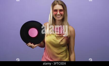 Blonde Frau hält mit der Hand eine Vinylplatte, lächelt und trägt eine rosa Sonnenbrille und ein drapiertes Oberteil im Studio; fröhliche Nostalgie Retro-Musik. Stockfoto