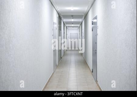 Leerer moderner Wohnkorridor mit weißen Wänden, grauen Türen, gefliestem Boden und hellen Deckenleuchten, in Perspektive betrachtet Stockfoto