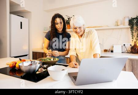 Zwei Frauen nutzen einen Laptop, während sie in einer modernen Hausküche einen frischen Salat zubereiten Stockfoto