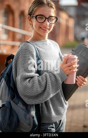Junge Frau hält einen Laptop und einen wiederverwendbaren Becher und trägt einen grauen Pullover, während sie tagsüber im Freien spaziert Stockfoto