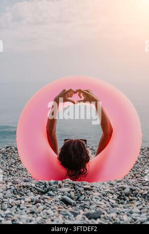 Frau Strand Herz Hände, die ein Liebessymbol bilden, eingerahmt von einem rosa aufblasbaren Schlauch an einem sonnigen Kieselstrand. Stockfoto