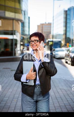 Eine lächelnde reife Geschäftsfrau spricht mit ihrem Smartphone, während sie einen Laptop auf einer Straße in Helsinki vor einem modernen Bürogebäude hält. Stockfoto
