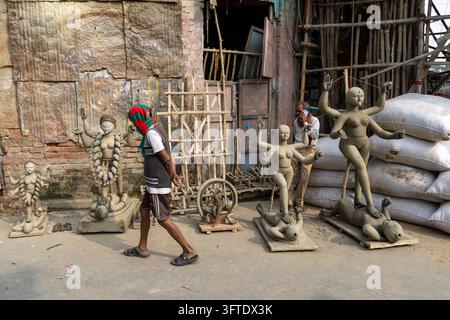 Walker mit einer grün-roten Kopfbedeckung geht an rohen Ton-Göttinnenstatuen auf Holzsockeln im Bezirk Kumartuli, Westbengalen, Kalkutta, Indien vorbei Stockfoto