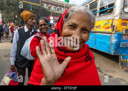 Eine ältere Frau mit grauen Haaren und einem roten Kopftuch lächelt und hebt ihre offene Hand in Richtung der Kamera in der Nähe von Bussen, West Bengal, Kalkutta, Indien Stockfoto