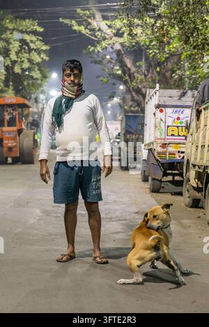 Ein Mann in einem weißen Sweatshirt mit einem Schal über dem Gesicht steht auf einer ruhigen Straße mit geparkten Lastwagen und einem braunen Hund, West Bengal, Kalkutta, Indien Stockfoto
