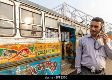 Ein Mann in einem leichten Hemd hält ein Telefon ans Ohr neben einem alten verzierten Bus unter einer großen Stahlbrücke, West Bengal, Kalkutta, Indien Stockfoto