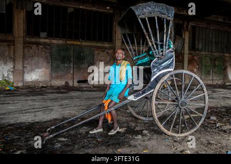 Ein Mann in einer türkisfarbenen Tunika sitzt am Rand einer schwarzen von Hand gezogenen Rikscha mit nach hinten geneigtem Kopf in einer Halle, West Bengal, Kalkutta, Indien Stockfoto