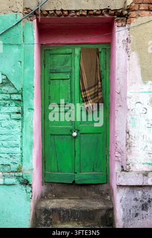 Eine mit einem Vorhängeschloss gesicherte grüne Holztür in einer rosa gestrichenen Nische in einer mintgrünen Wand mit einem karierten Tuch, das darüber hängt, West Bengal, Kolkata, Indien Stockfoto