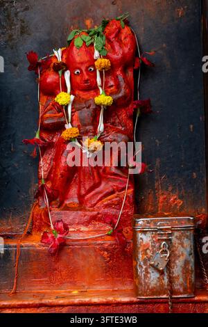 Mit roter Farbe beschmierte Affengottfigur, geschmückt mit Ringelblumen und Hibiskusblüten, neben einer verschlossenen Opferdose, West Bengal, Kalkutta, Indien Stockfoto