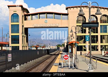 Eine braune Fußgängerbrücke mit der Aufschrift San Bernardino überspannt Bahngleise zwischen Stationsgebäuden, mit Plattformen, kunstvollen Laternen und Bergen darunter, weiß getüncht Stockfoto