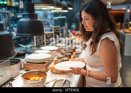 Frau wählt warmes Essen, während sie einen Teller an einem Hotel-Frühstücksbuffet hält. Morgenszenebild repräsentiert Selbstbedienung und Gastfreundschaft für Reisende Stockfoto