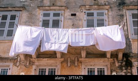 Weiße Laken trocknen an einer Wäscheleine über einer schmalen Gasse, eingerahmt von verwitterten Fensterläden und alten Steinbalkonen in der Altstadt von Korfu, Griechenland Stockfoto