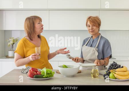 Zwei Frauen sind in einer Küche und bereiten einen Salat zu. Eine Frau hält ein Glas und spricht, während die andere bereit ist, den Salat zu servieren. Frische Früchte und Gemüse Stockfoto