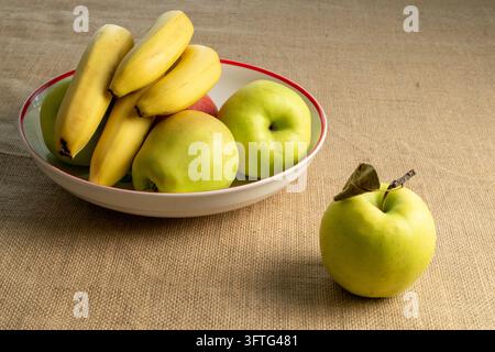 Frische gelbe Bananen und reife grüne und rote Äpfel in einer Keramikschale auf einem rustikalen Sackleinenhintergrund. Stockfoto