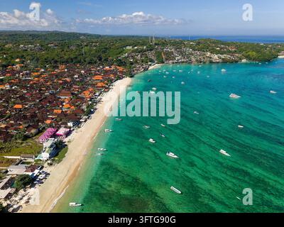 Boote, die über klarem türkisem flachem Wasser entlang eines geschwungenen Sandstrands vertäut sind, grüne bewaldete Hügel und eine Küstensiedlung im Hintergrund, Nusa Lembongan Stockfoto