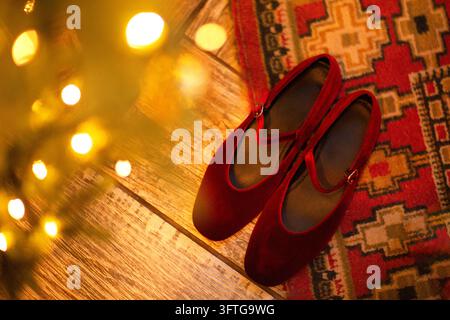 Rote Ballettschuhe ruhen neben glühenden Lichterketten auf einem Holzboden, teilweise überlappend einen farbenfrohen gemusterten Teppich in einer warmen Innenszene. Dopamin Stockfoto