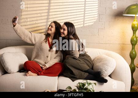 Zwei glückliche Freundinnen lächeln und machen ein Selfie auf einem Sofa in einem Wohnzimmer Stockfoto