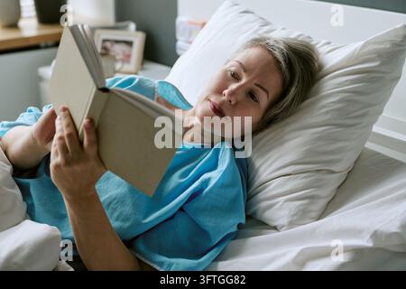 Mittlere Altersfrau liest ein Buch, während sie auf einer medizinischen Station ruht Stockfoto