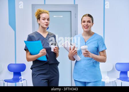 Konzept der medizinischen Ausbildung, des Praktikums und der Zusammenarbeit im Gesundheitswesen: junge Ärztinnen oder Praktikantinnen in OP-Kleidung, die während der Pause Ordner und Kaffee halten Stockfoto