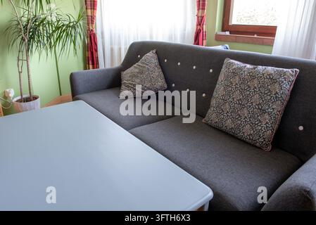 Ein graues gepolstertes Sofa mit floral gemusterten Kissen steht neben einem blauen Tisch in einem gemütlichen Wohnzimmer, mit einer Topfpflanze, durchsichtigen Vorhängen und einem Stockfoto