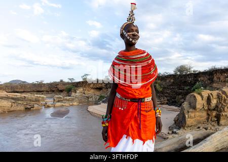 Frau des Samburu-Stammes in einem orangefarbenen Kleid mit einem perlenbesetzten Gesichtsband und riesigem rotem Perlenkragen am Ufer eines Flusses, Samburu County, Samburu, Kenia Stockfoto