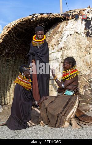 Drei Frauen des Turkana-Stammes, die in Perlenhalsketten vor einer traditionellen, mit Leder bedeckten Hütte in Marsabit County, Loiyangalani, Kenia, plaudern und lächeln Stockfoto
