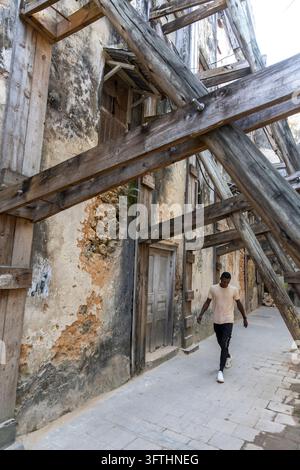 Junger Mann geht eine Gasse entlang unter hölzernen Stützbalken, die zerbrochene Korallensteinhäuser mit bröckelnden Wänden stützen, Stone Town, Tansania Stockfoto