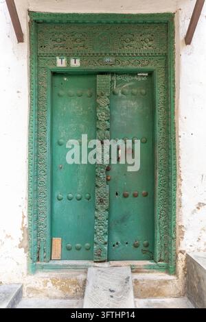 Grüne antike geschnitzte Holztür mit Messingnieten und einem verzierten Blumenrahmen sowie Kreidebeschriftung in einer weiß getünchten Wand, Stone Town, Tansania Stockfoto
