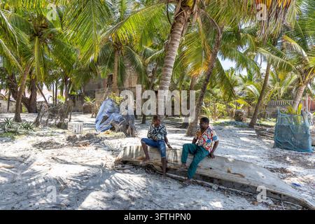 Zwei Männer unterhalten sich auf einem alten hölzernen Kanu-Rumpf unter Kokospalmen an einem sandigen Strand mit Fischernetzen und einfachen Hütten, Matemwe, Tansania Stockfoto