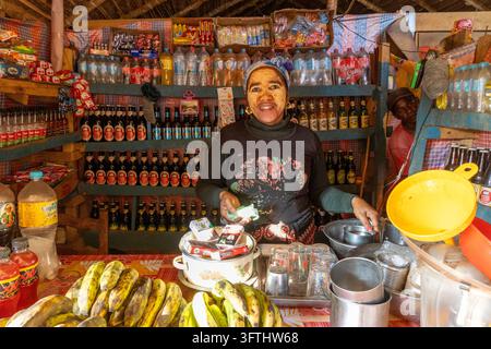 Frau, die eine traditionelle gelbe Gesichtsmaske trägt, hinter der Theke eines kleinen Ladens, der mit Getränken und Bananen bestückt ist, Menabe, Tsiribihina, Madagaskar Stockfoto