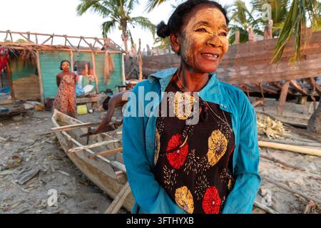 Lächelnde Vezo-Frau, die eine traditionelle Gesichtsmaskenpaste trägt, während sie in der Nähe von Holzbooten steht, die unter Konstruktion sind, Menabe, Belo sur Mer, Madagaskar Stockfoto