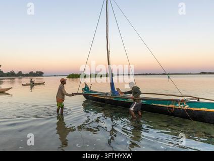 Zwei Vezo-Fischer waten in flachem Wasser und bereiten ein Ausrigger-Segelboot bei Sonnenuntergang vor, während ein anderer vorbeerpaddelt, Menabe, Belo sur Mer, Madagaskar Stockfoto