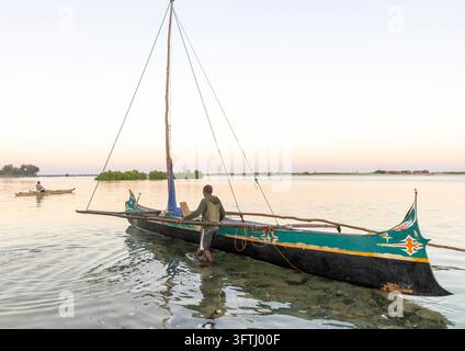 Ein Vezo-Fischer watet ins Wasser und schiebt ein dekoriertes Ausrigger-Segelboot bei Sonnenuntergang aufs Meer, Menabe, Belo sur Mer, Madagaskar Stockfoto
