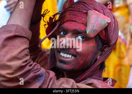 Nahaufnahme eines lächelnden jungen Mannes mit rotem Pulver im Gesicht, der von unter einem kastanienbraunen Turban bei Lathmar Holi in Uttar Pradesh, Barsana, Indien aufblickt Stockfoto