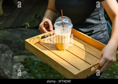Ein hölzernes Tablett mit Eiskaffeegetränk, Hand, hölzernes Tablett mit Eiskaffeegetränk im Freien, mit einer Person, die das Getränk neben einem schattigen Gartengraben trägt und Stockfoto