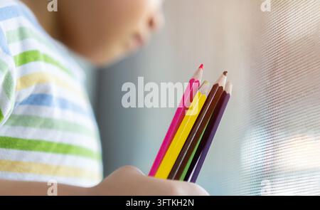 Ein Junge, ein Kind, das verschiedene bunte Stifte in verschiedenen Farben auf dem Hintergrund eines Klassenzimmerfensters hält, Nahaufnahme, Kinder malen und Hobbykonzept, Kopierraum Stockfoto